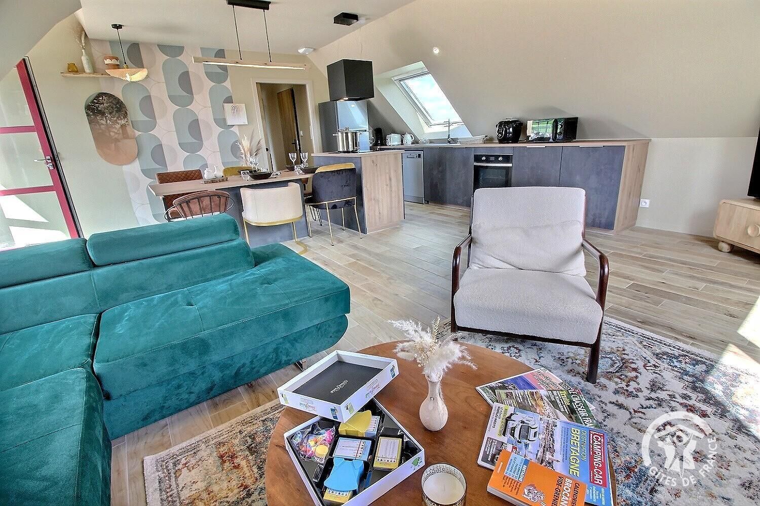 A modern living room with a teal sofa, a grey armchair, and a coffee table with magazines and a candle. A kitchen with a skylight and a dining area with a table and chairs are visible in the background.