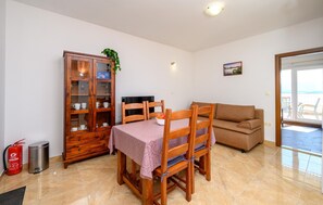 Uma área de refeições com uma mesa e cadeiras de madeira, um armário com portas de vidro, um sofá e vista para o mar através de uma porta de vidro.
