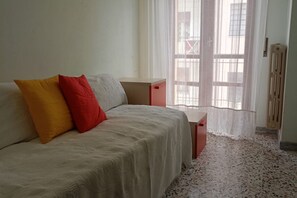 Una cama con cubierta blanca, una almohada roja y otra amarilla, una mesita de noche de madera y un radiador.
