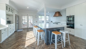 A modern kitchen with white cabinetry, a central island with stools, a large oven, and a view to the outside. - The Grand on Macquarie | Pool & Spa, Ultimate Getaway (Dubbo)