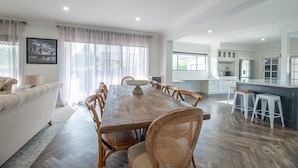 A spacious living room with a large wooden dining table, comfortable seating, and a view of the kitchen area. - The Grand on Macquarie | Pool & Spa, Ultimate Getaway (Dubbo)