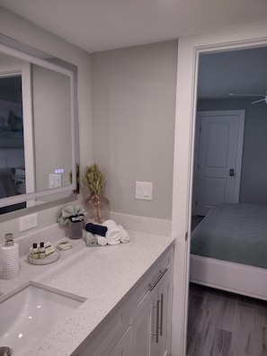 A modern bathroom with a white speckled countertop, a large mirror, and a bed visible through an open door. - Luxury Beach Retreat Steps from Barefoot Beach & Restaurants (Bonita Springs)