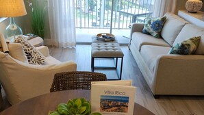 Una sala de estar con sofá, sillones, una mesa de comedor con planta y un balcón con vistas.