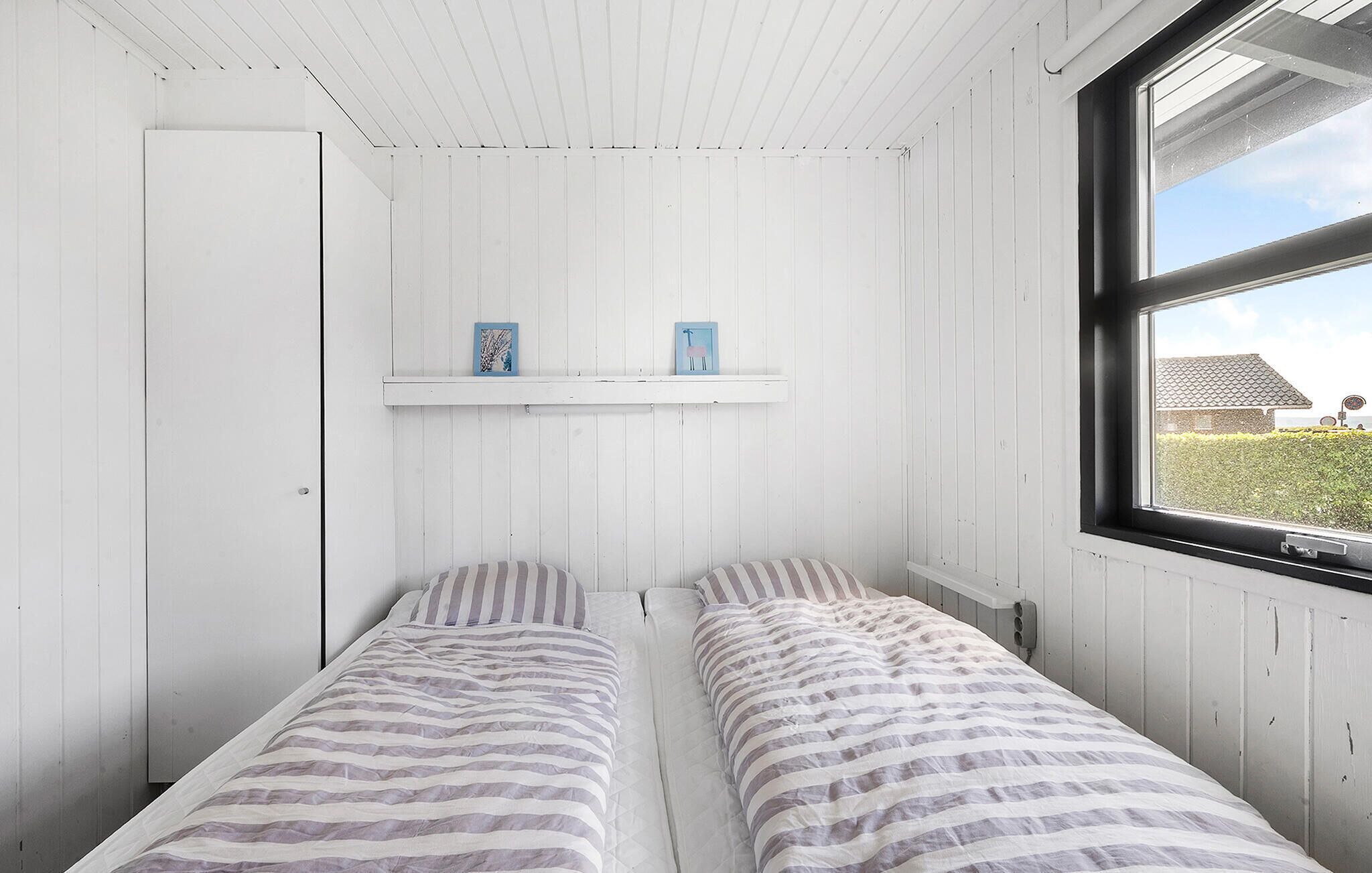 Un alojamiento pequeño y de una sola habitación con dos camas individuales, paredes de madera blanca, un ventanal con vista a un edificio y una puerta.