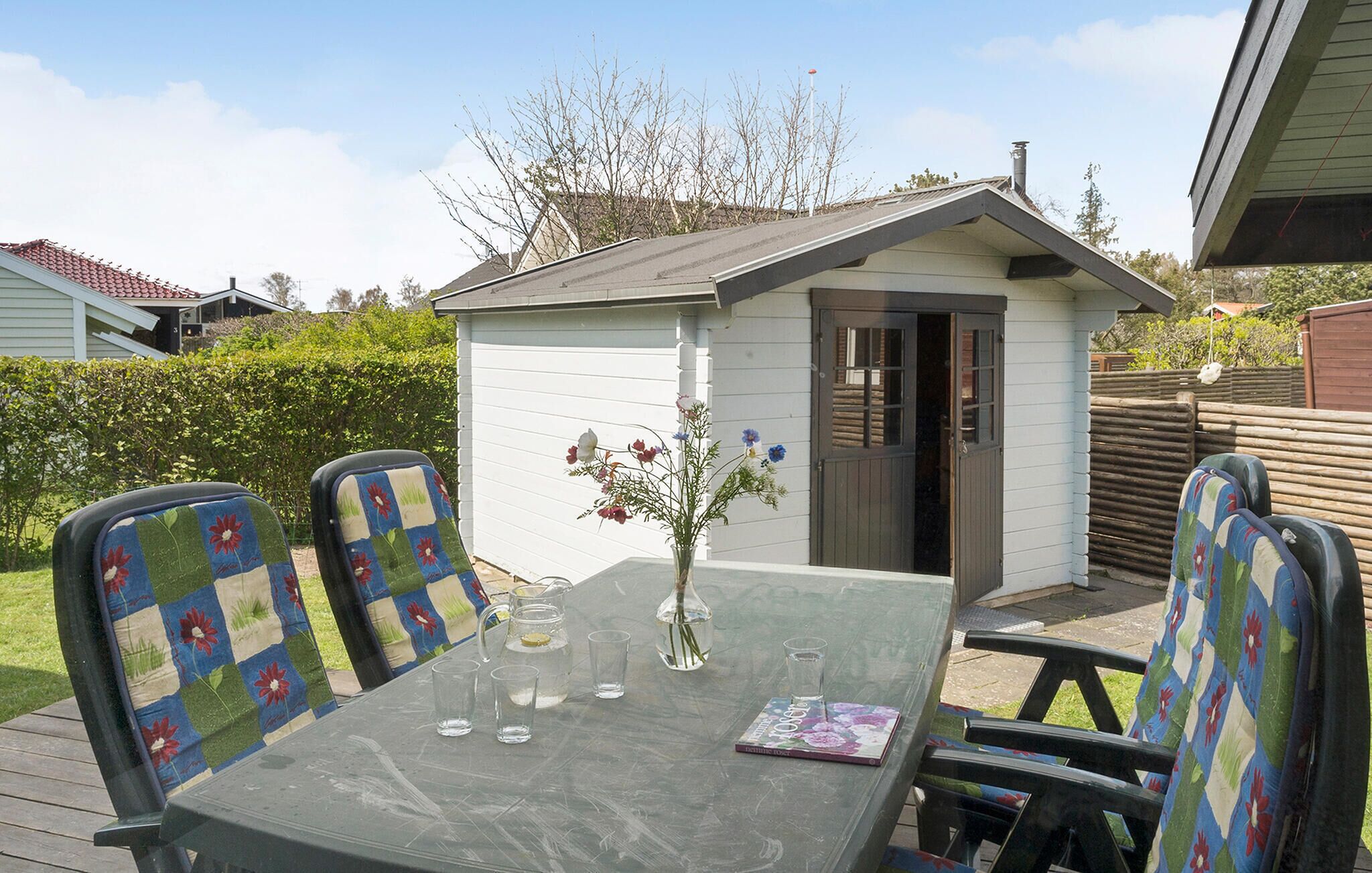 A patio with a glass table, four chairs with floral upholstery, a small shed, and a vase with flowers.