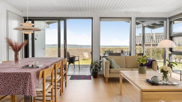 Ein großzügiger Wohnbereich mit einem großen Tisch, Stühlen, einer Couch und einem Couchtisch. Große Fenster bieten einen Blick ins Freie.