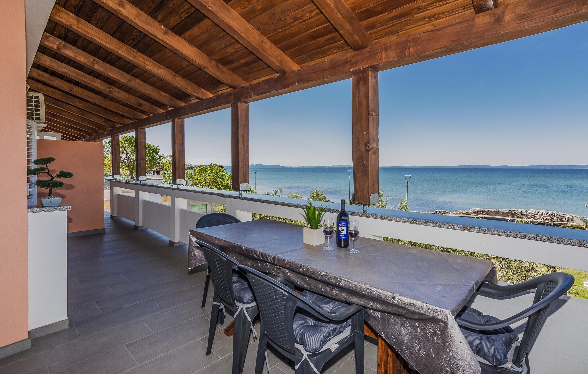 An outdoor patio with a wooden ceiling, a table with chairs, a bottle of wine, and a view of the sea.