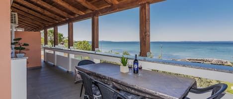 An outdoor patio with a wooden ceiling, a table with chairs, a bottle of wine, and a view of the sea.