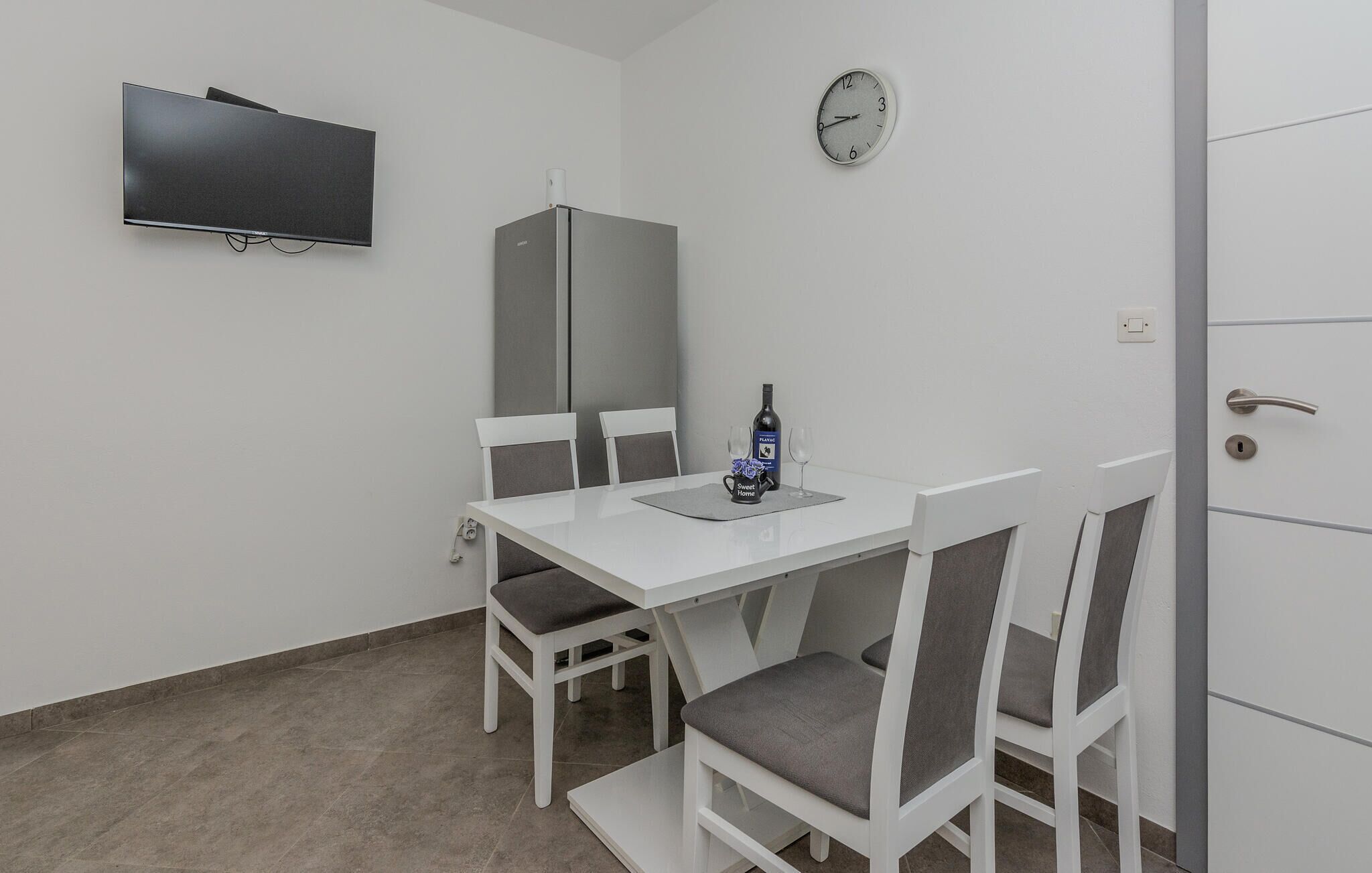 A small, modern room with a white table and chairs, a flat-screen TV mounted on the wall, a refrigerator, and a clock.