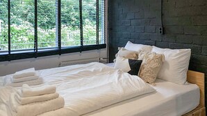 Un lit bien fait, avec du linge de lit et des serviettes blanches, un mur en briques sombres et une fenêtre donnant sur un paysage verdoyant.
