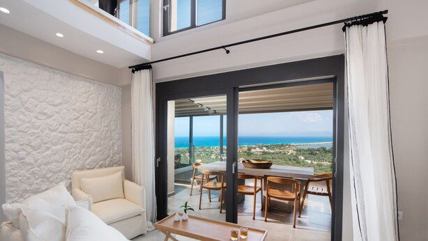 Een moderne woonkamer met een witte bank, een houten salontafel en uitzicht op zee door grote glazen deuren.
