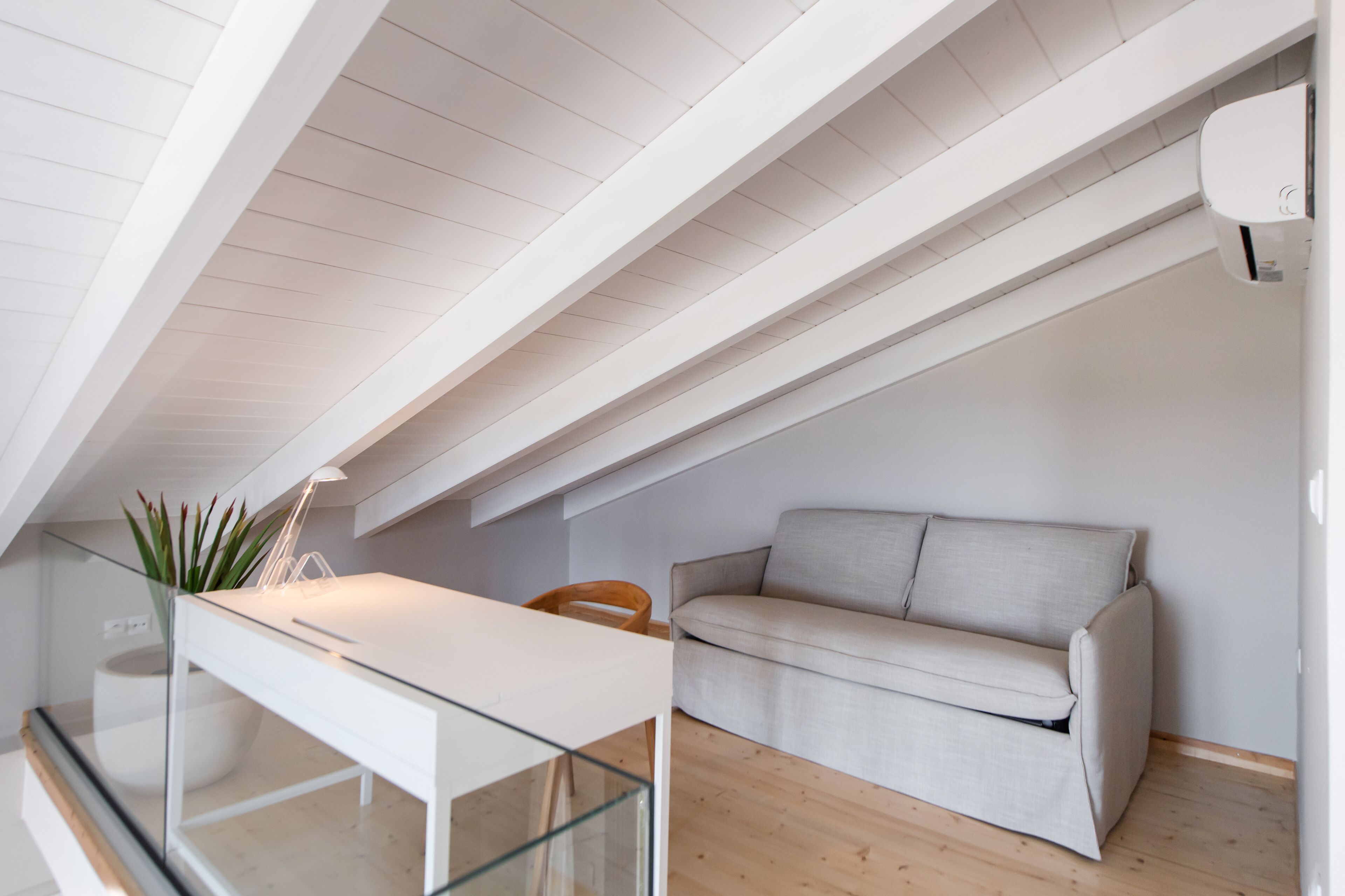 A modern loft bedroom with a glass dining table, a beige sofa, and a white ceiling with exposed beams.