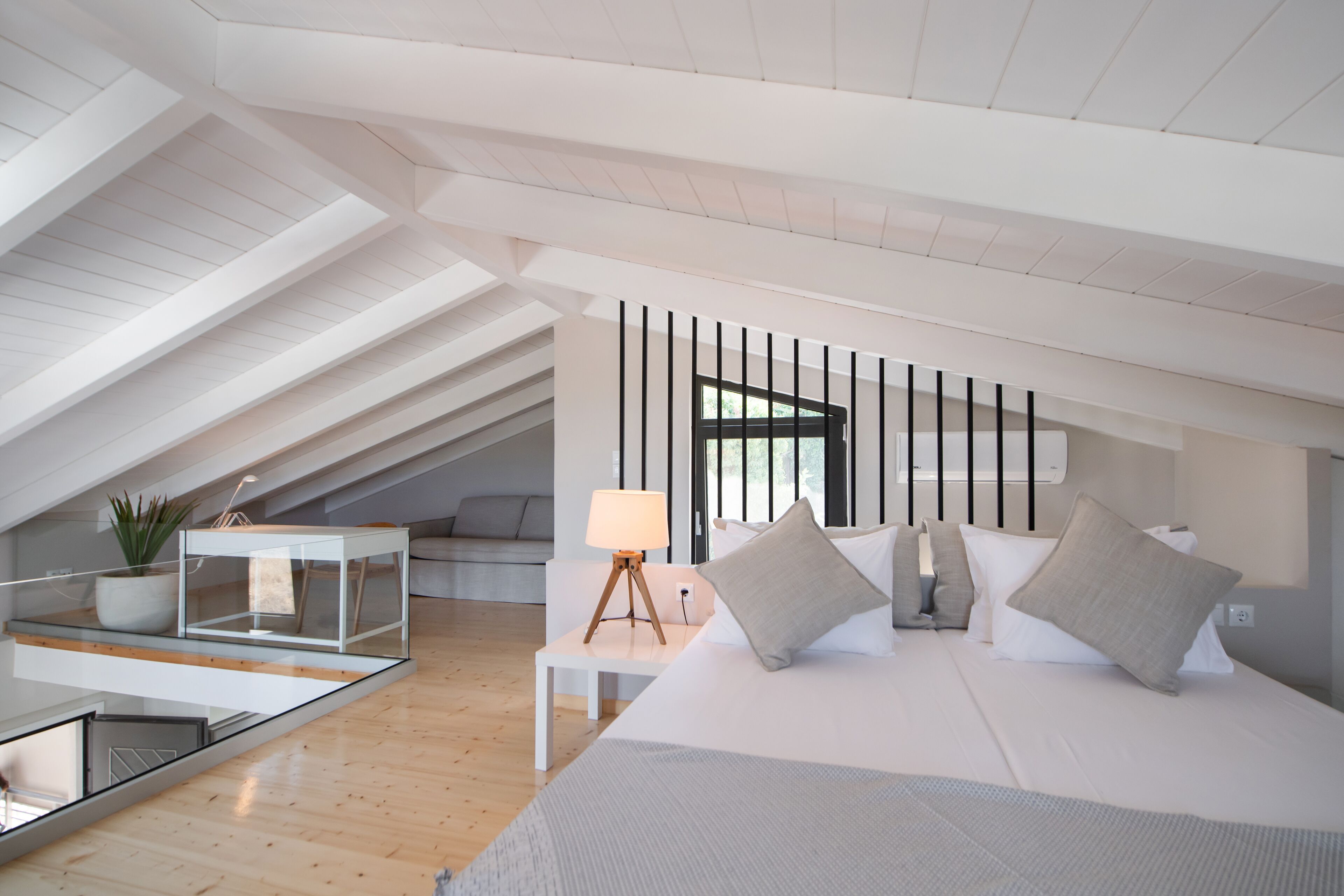 A modern bedroom with a large bed, a bedside table with a lamp, a small table with a plant, and a view of the outdoors through a window.