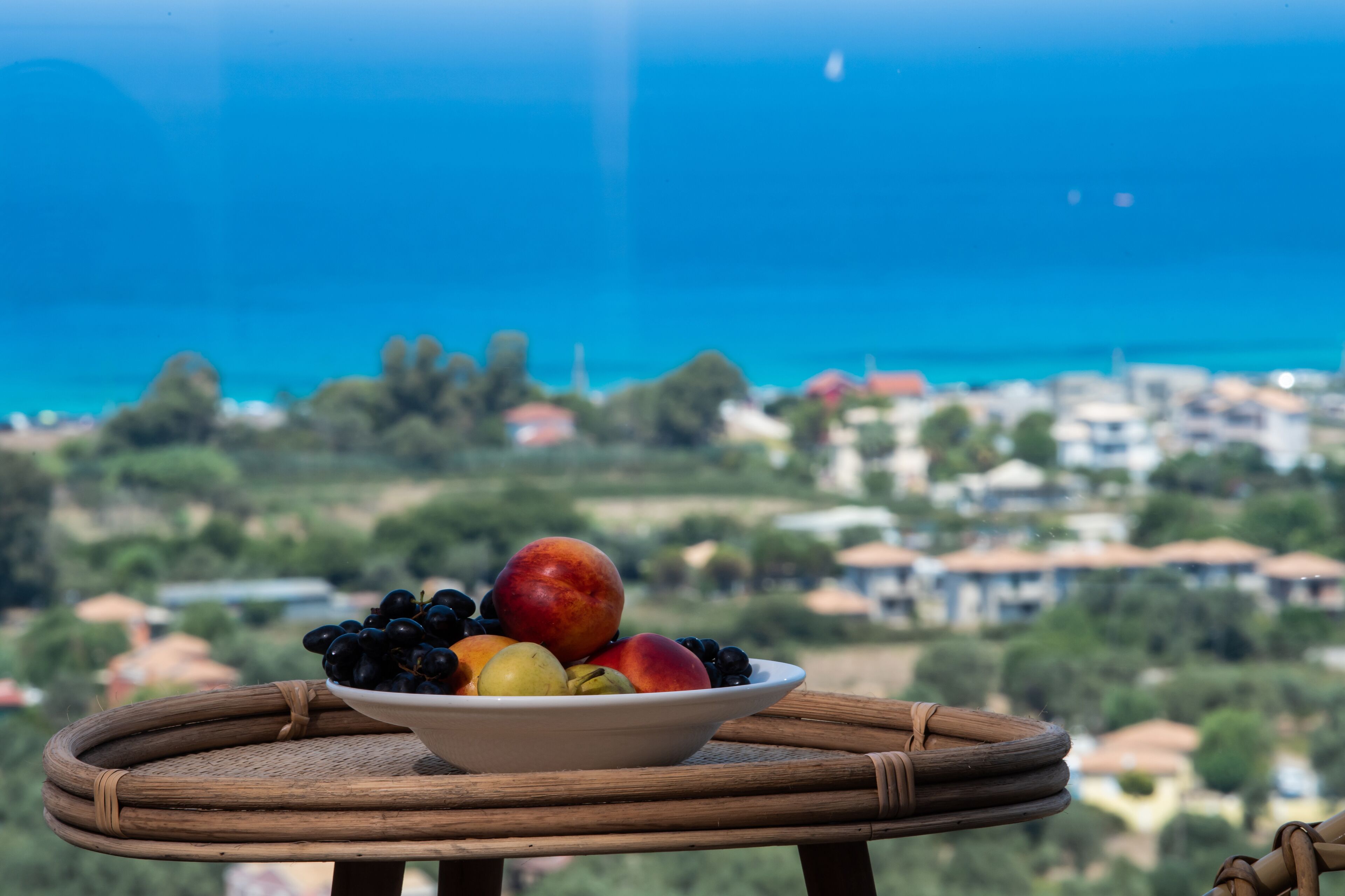 Un panier de fruits est posé sur une table ronde en osier, avec une vue sur la côte en arrière-plan.