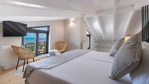Een slaapkamer met een groot bed, een flatscreen-tv aan de muur, een rieten stoel en uitzicht op de zee door een groot raam.