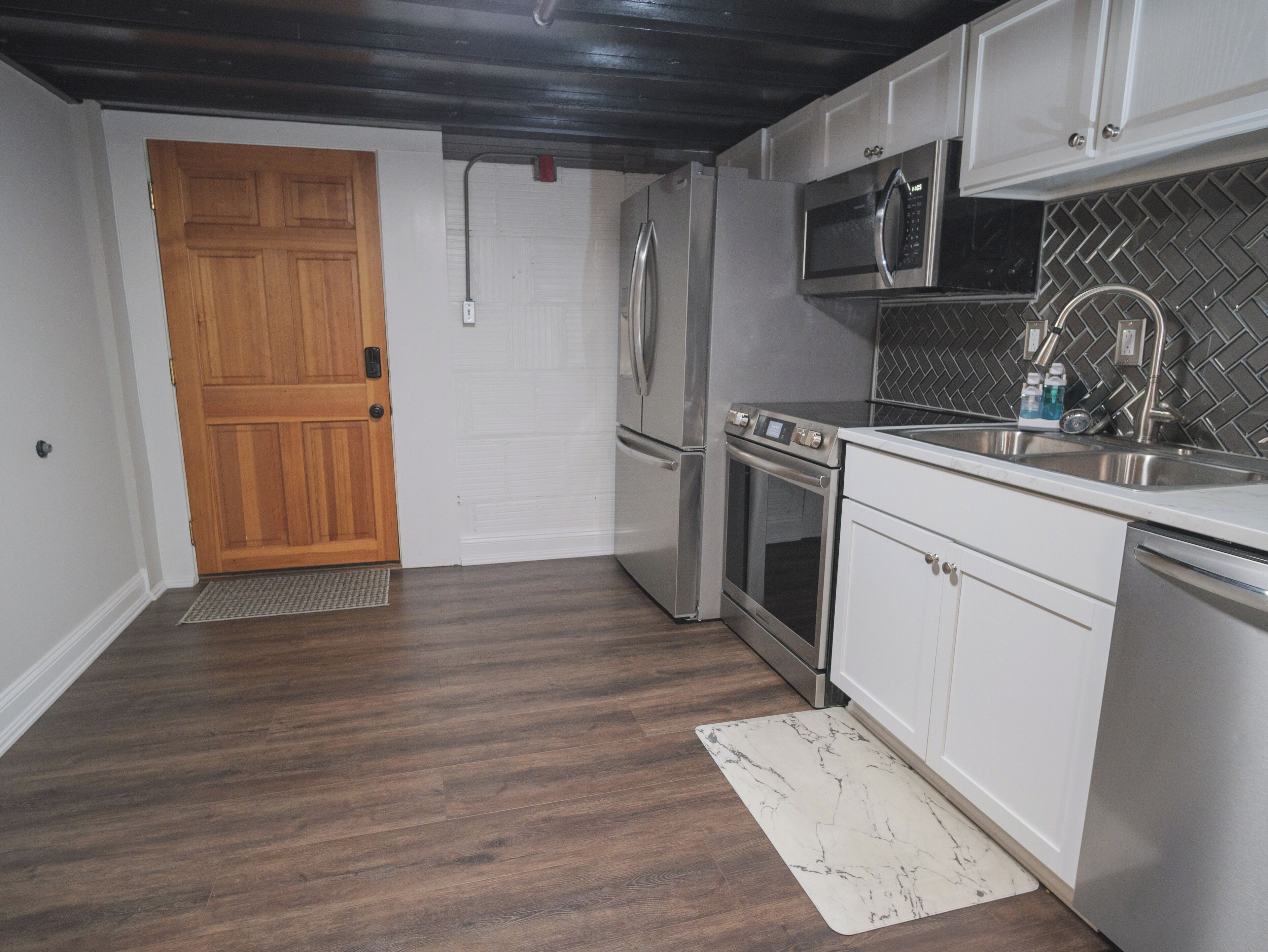 Una cocina con armarios blancos, electrodomésticos de acero inoxidable, una puerta de madera y un salpicadero de baldosas tipo espiga.