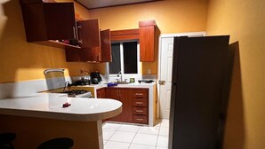 A compact kitchen with a white countertop, wooden cabinets, a refrigerator, and a window. - Casa de Vacaciones (La ceiba)