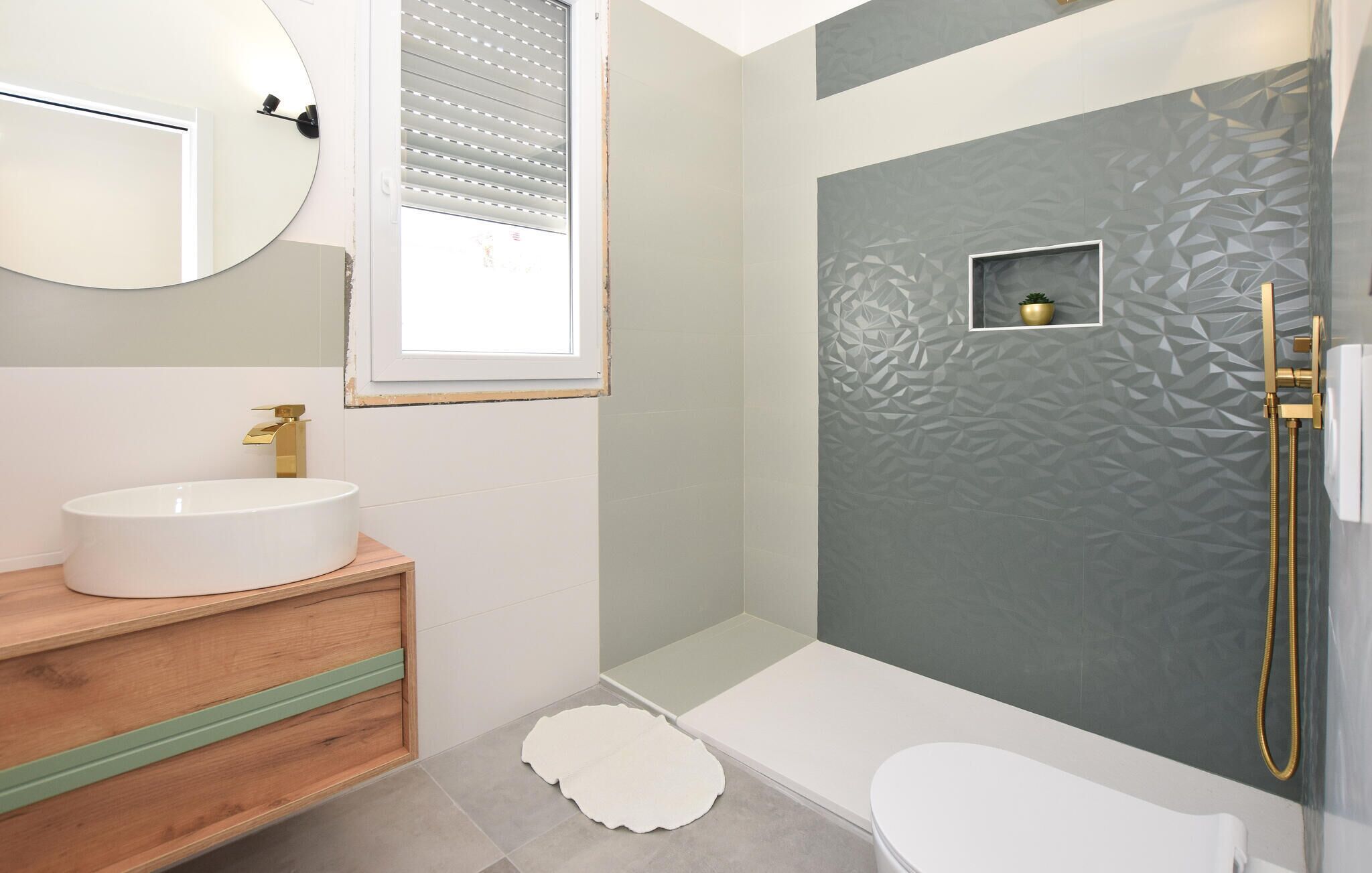 Ein Badezimmer mit rundem Spiegel, weißem Waschbecken, Toilette und Duschbereich mit strukturierter Wand.