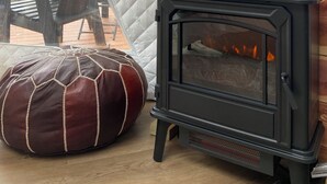 A cozy room with a quilted wall, a black wood-burning stove, a white vase with dried plants, and a brown leather pouf. - RIVER VIEW Cliff Dome Glamping @ White River, minutes to fishing, hikes! (Cotter)