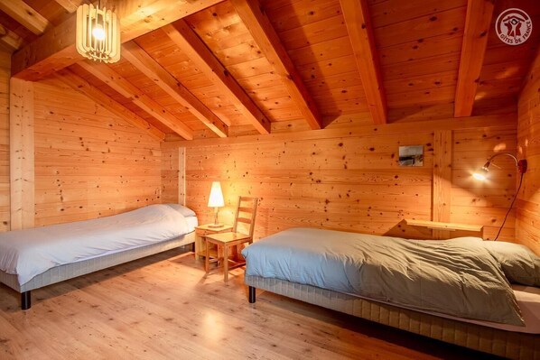 A cozy wooden cabin room with two single beds, a small table with a lamp, and a slanted ceiling.