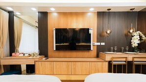 Une chambre d’hôtel dotée d’un grand téléviseur à écran plat fixé à un mur en bois, d’un bureau en bois avec une chaise, d’un lit avec une literie blanche et d’une petite table sur laquelle est posé un vase de fleurs.