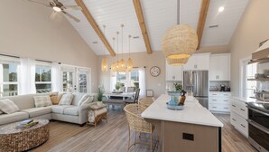 A spacious living room with a large sofa, a dining area with wicker chairs, a kitchen with white cabinets and stainless steel appliances, and a ceiling fan.