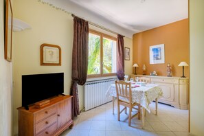 A room with a TV, wooden cabinet, dining table with chairs, and a window with curtains. - Apartment 'Grand Studio Bougainvillée' with Shared Pool, Private Terrace and Wi-Fi (Cagnes-sur-Mer)