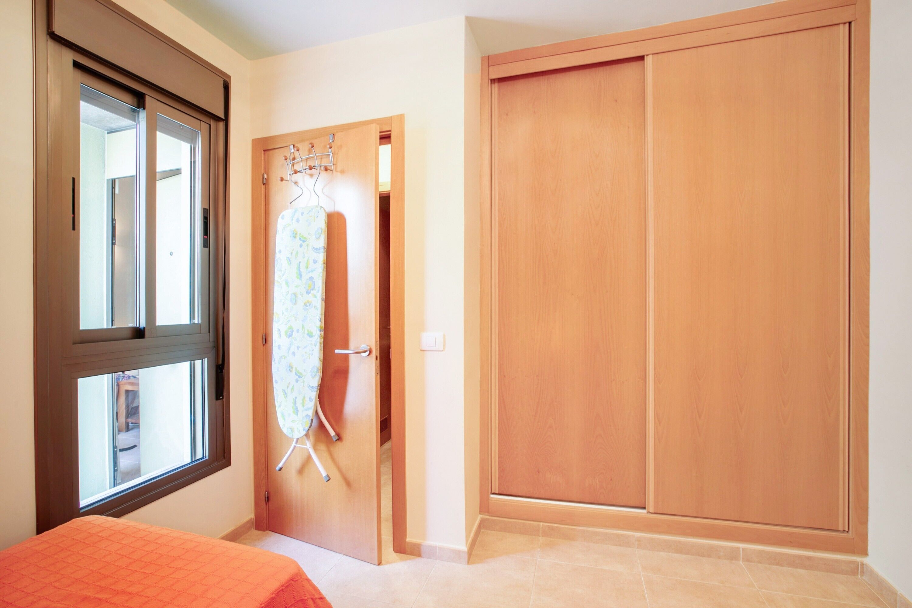 A bedroom with a bed, a window, a wooden wardrobe, and a door with a hanging rack.