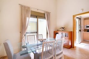 A dining area with a glass table, white chairs, a wooden cabinet, and a view of the kitchen.