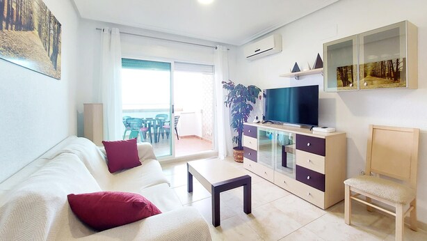 Una sala de estar con sofá, una mesa de centro, televisor sobre un mueble, una silla y un balcón con sillas y una mesa.