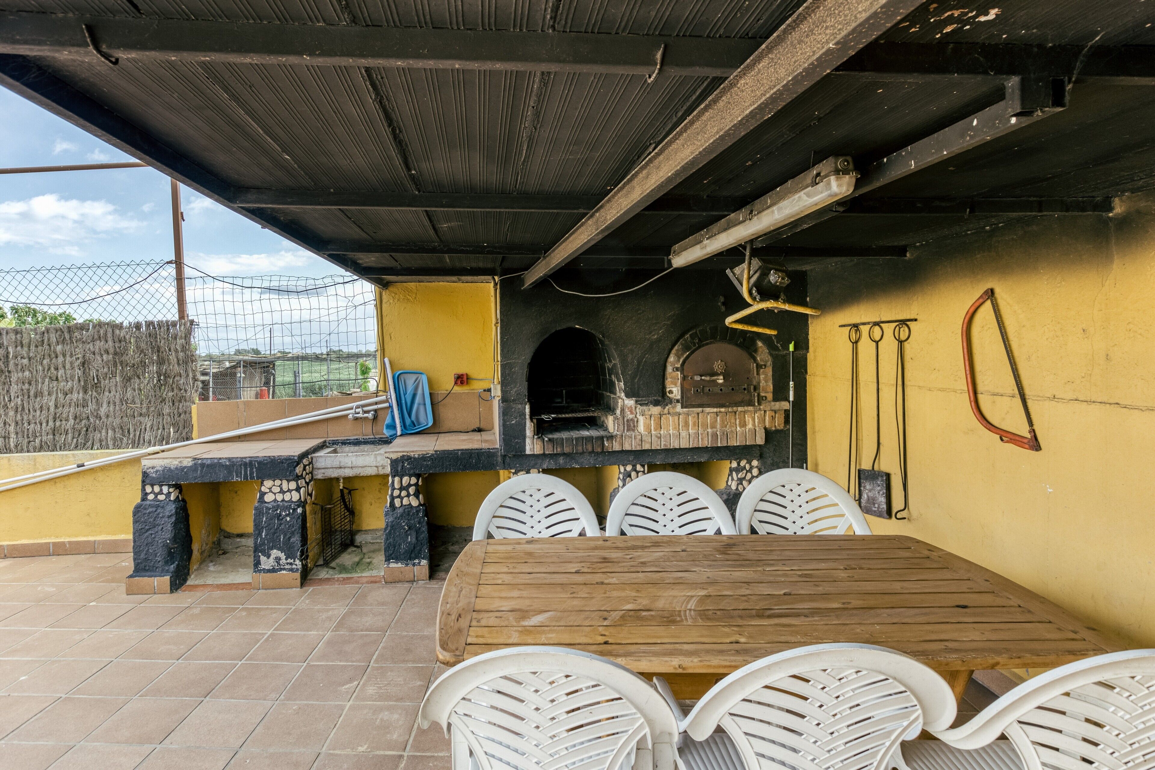 Un patio al aire libre con una mesa de madera y sillas blancas, un horno de ladrillo y una pared amarilla.