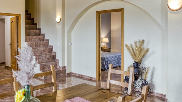 Un coin repas avec une table et des chaises en bois, un vase contenant des plantes séchées, un escalier et un lit recouvert d’une couvre-lit à motifs bleus.