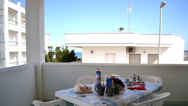 Un balcón con una mesa lista para el desayuno, sillas blancas y vista a un edificio con balcones y cielo despejado.