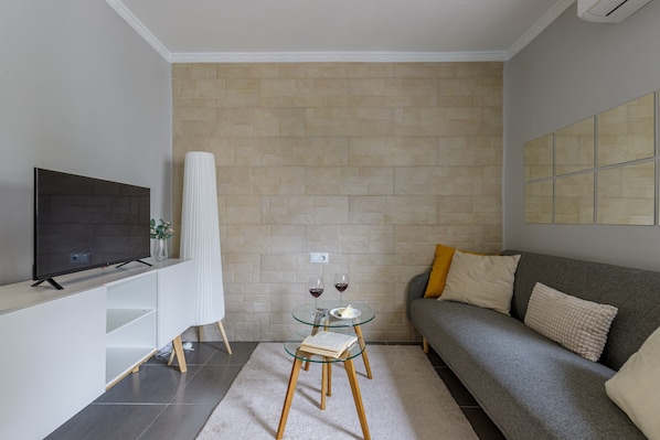 Un salon moderne avec un téléviseur à écran plat, une table en verre avec deux verres à vin, un canapé avec des coussins et un mur recouvert de carreaux.
