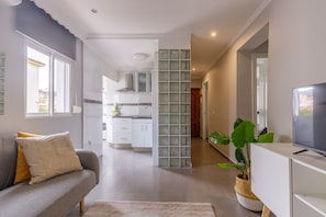 Un salon moderne avec un canapé gris, un téléviseur à écran plat, une grande plante verte et une cuisine en arrière-plan.