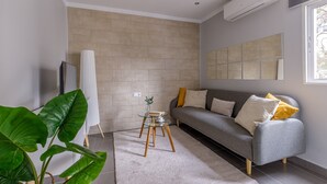Una sala de estar moderna con un sofá gris, una mesa de centro de vidrio, una planta verde grande y una lámpara blanca.