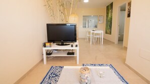 Una sala de estar con un televisor sobre un mueble bajo, una zona de comedor con una mesa y sillas blancas, y un pasillo que lleva a un baño.