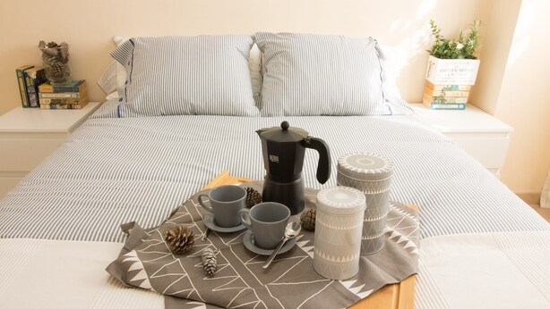 Una cama bien hecha con ropa de cama a rayas, una cafetera, tazas y una bandeja con servilletas de estampado geométrico.