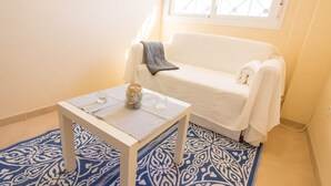 Una habitación pequeña con un sofá blanco, un tapete azul estampado, una mesita con un tarro y vasos, y una ventana con diseño de cuadrícula.