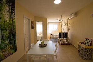 A dining area with a white table, chairs, and a painting of a forest. There is a white sofa and a TV in the background.