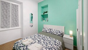 Un dormitorio con cama, mesita de noche, ventana con cortinas y estante en la pared con una planta.