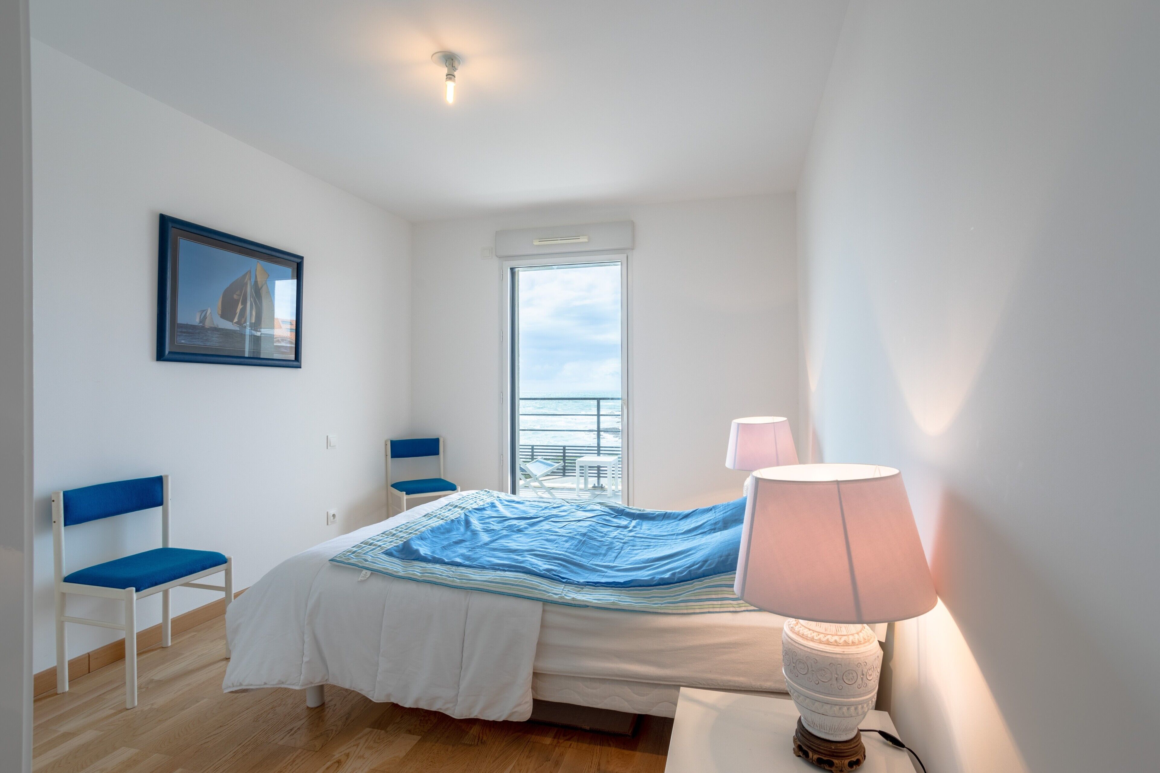 A bedroom with a bed, a chair, a window, a lamp, and a framed picture on the wall.