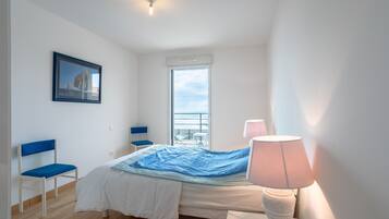 A bedroom with a bed, a chair, a window, a lamp, and a framed picture on the wall.