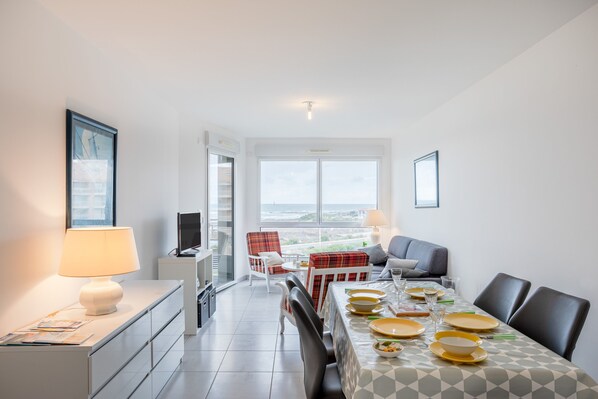 Un coin repas avec une table dressée pour quatre personnes, un canapé, un téléviseur et une fenêtre donnant sur un beau paysage.