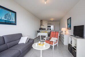 A modern living room with a grey sofa, a dining area with a table and chairs, a kitchen with a fridge, and a TV on a stand.