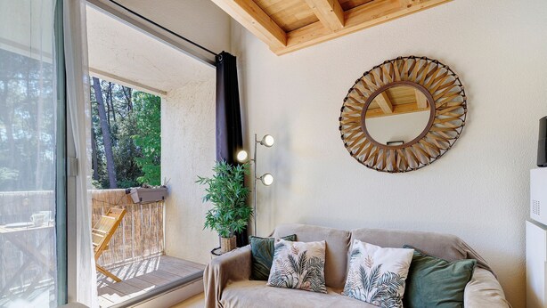 Ein gemütliches Zimmer mit einer gepolsterten Couch, einem runden Spiegel und Blick auf eine Holzterrasse und die grüne Umgebung.