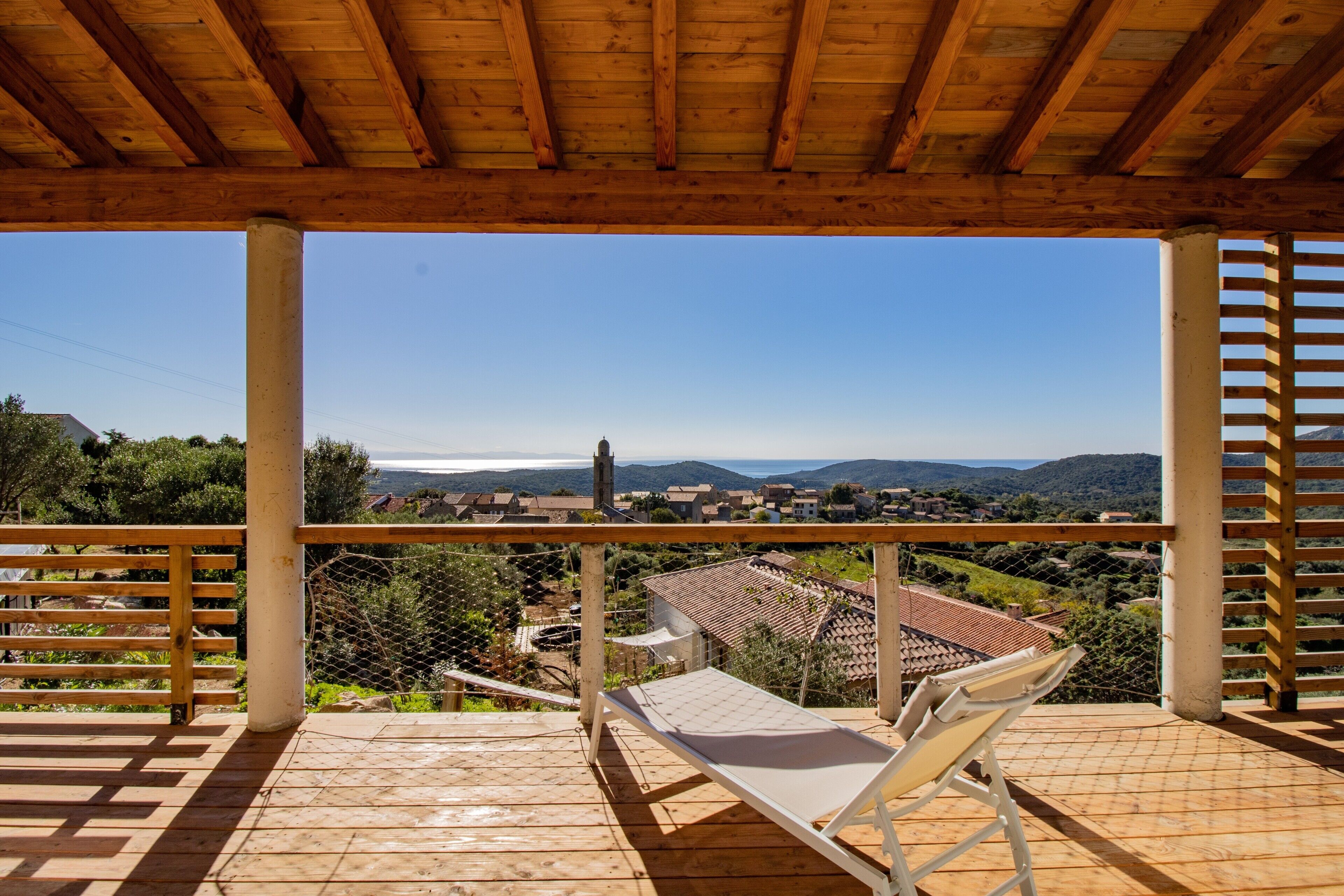 A wooden balcony with a white chair, overlooking a village with houses and hills.