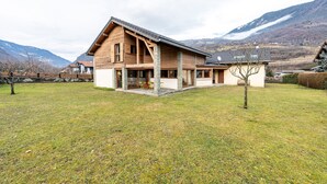 A wooden house with a sloped roof, stone pillars, and a grassy yard. - Chalet 'Pierre Et Bois' with Mountain View, Private Terrace and Wi-Fi (AIGUEBLANCHE)