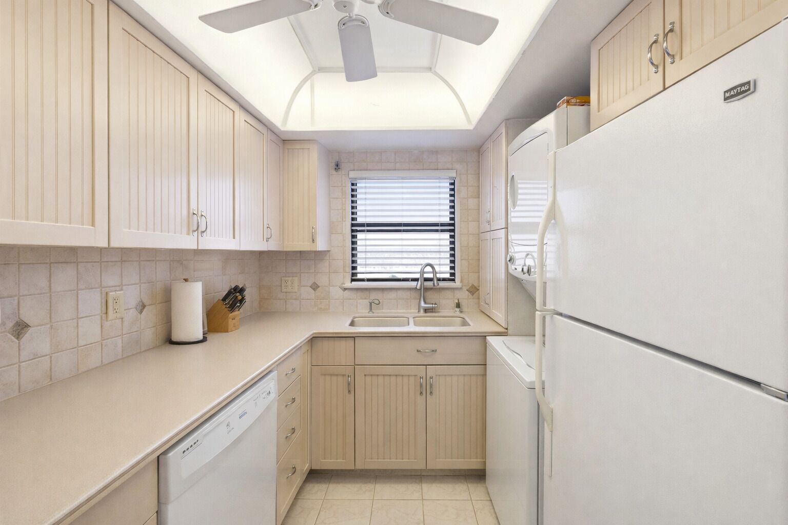 A compact kitchen with a dishwasher, a refrigerator, and a window with blinds.