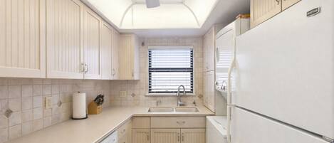 A compact kitchen with a dishwasher, a refrigerator, and a window with blinds.